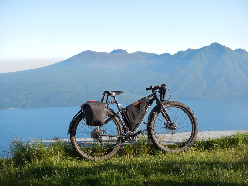 Right side view of a black Surly ECR bike with gear packs, on a grass hilltop, above a lake with mountains behind it