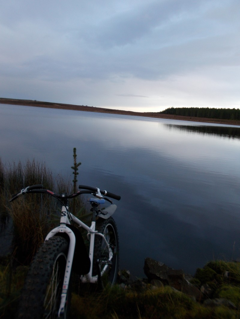 Front, left side view of a white Surly Pugsley fat bike, on a rocky grass bank, facing away from a calm lake