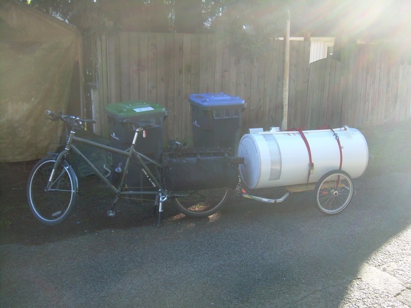 Left side view of an olive drab Surly Big Dummy bike with trailer loaded with a water heater, parked in an alley