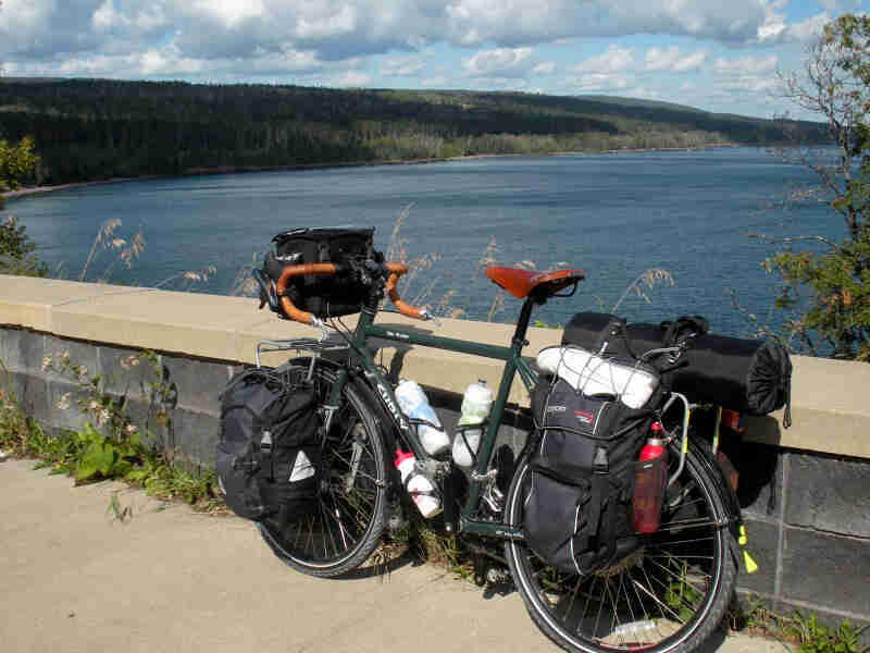 Left side view of a Surly Disc Trucker bike, green, alongside a block wall, with a lake and trees in the background