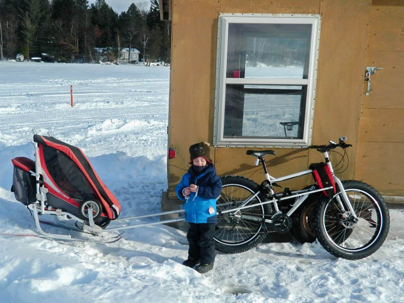 Front view of a child standing on a frozen lake in front of a Surly fat bike with ski trailer, next to a fish house