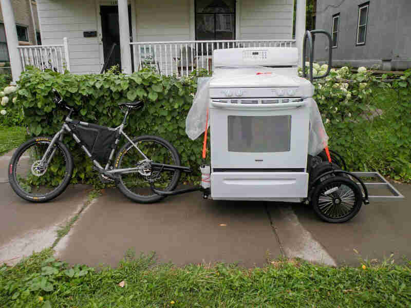 Left side view of a silver Surly bike with a trailer hauling a  stove attached, on a sidewalk in front of a home