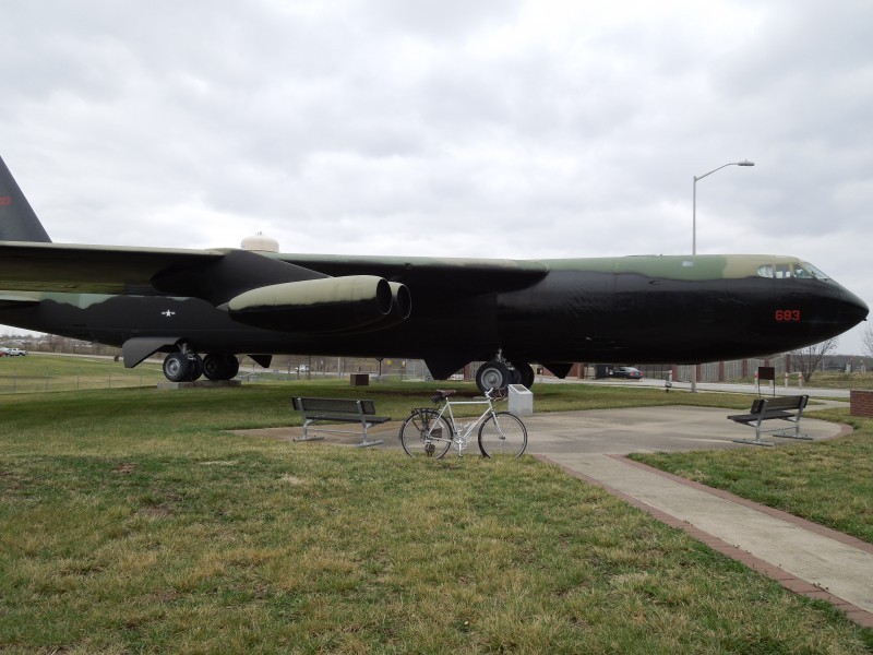 Right side view of a while Surly bike, parked in front of the right side of a B-52 bomber, in a grass field