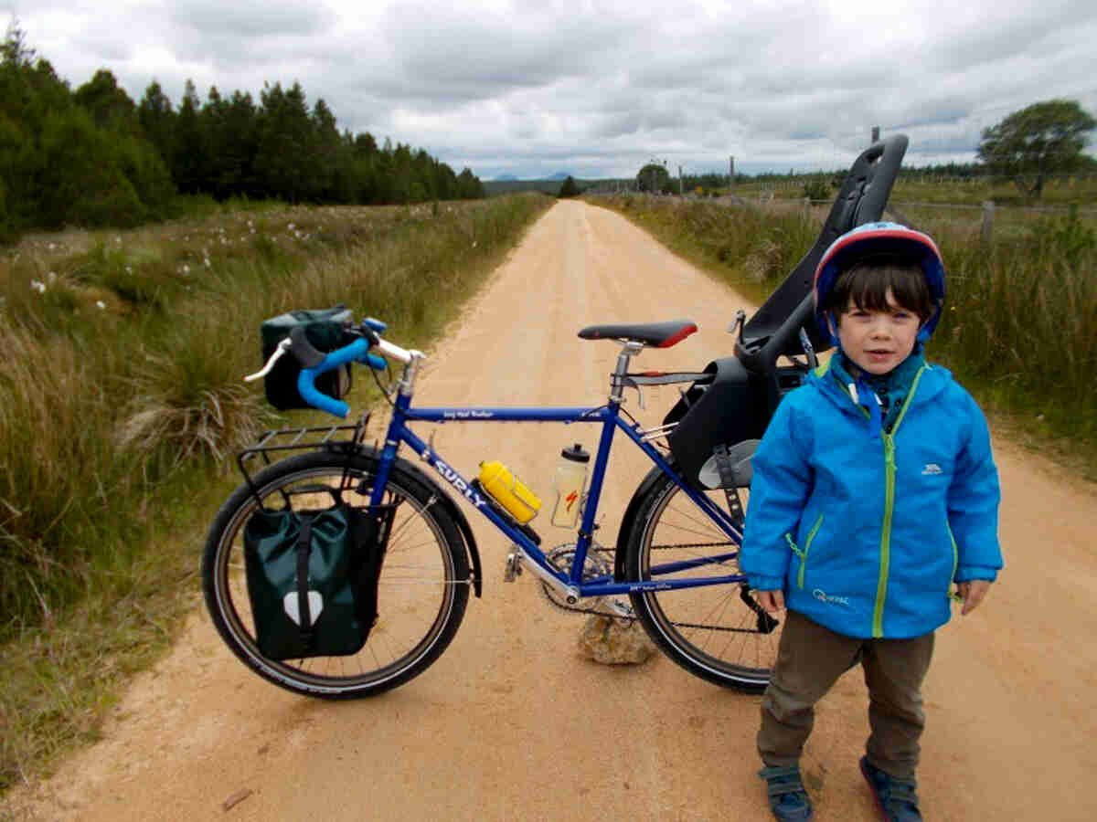 Left side view of a Surly Long Haul Trucker bike, parked on a gravel road, with a child standing near the rear wheel