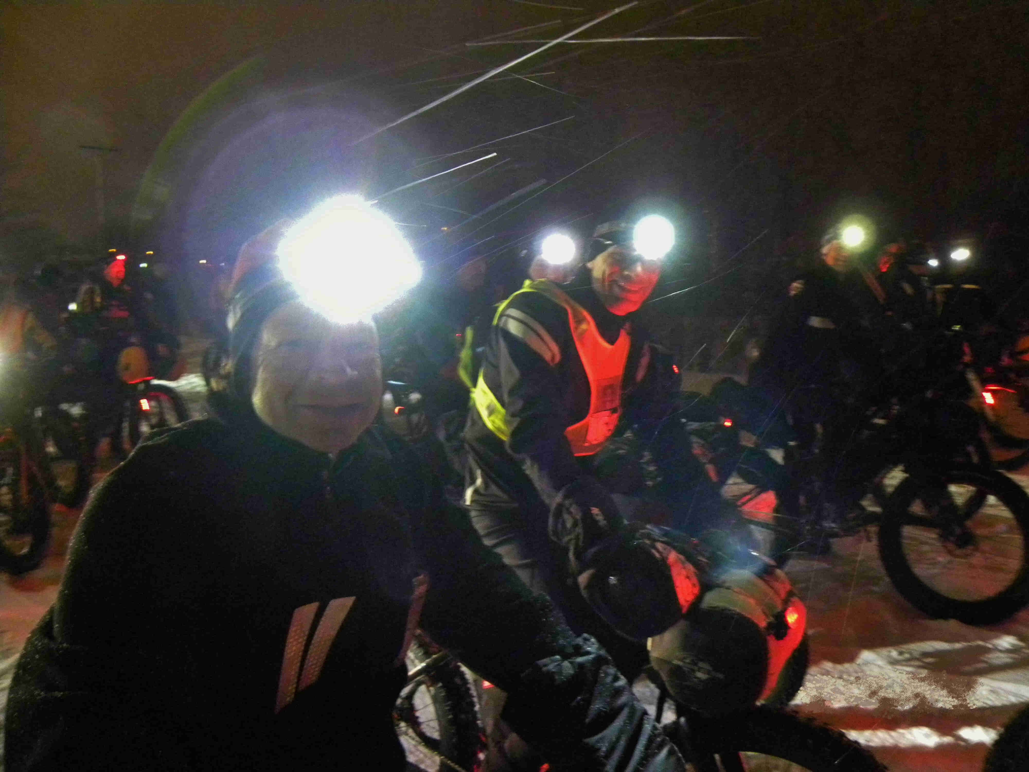 Side view of cyclists wearing winter outerwear and headlamps, lined up side by side on the bikes, at night in the snow