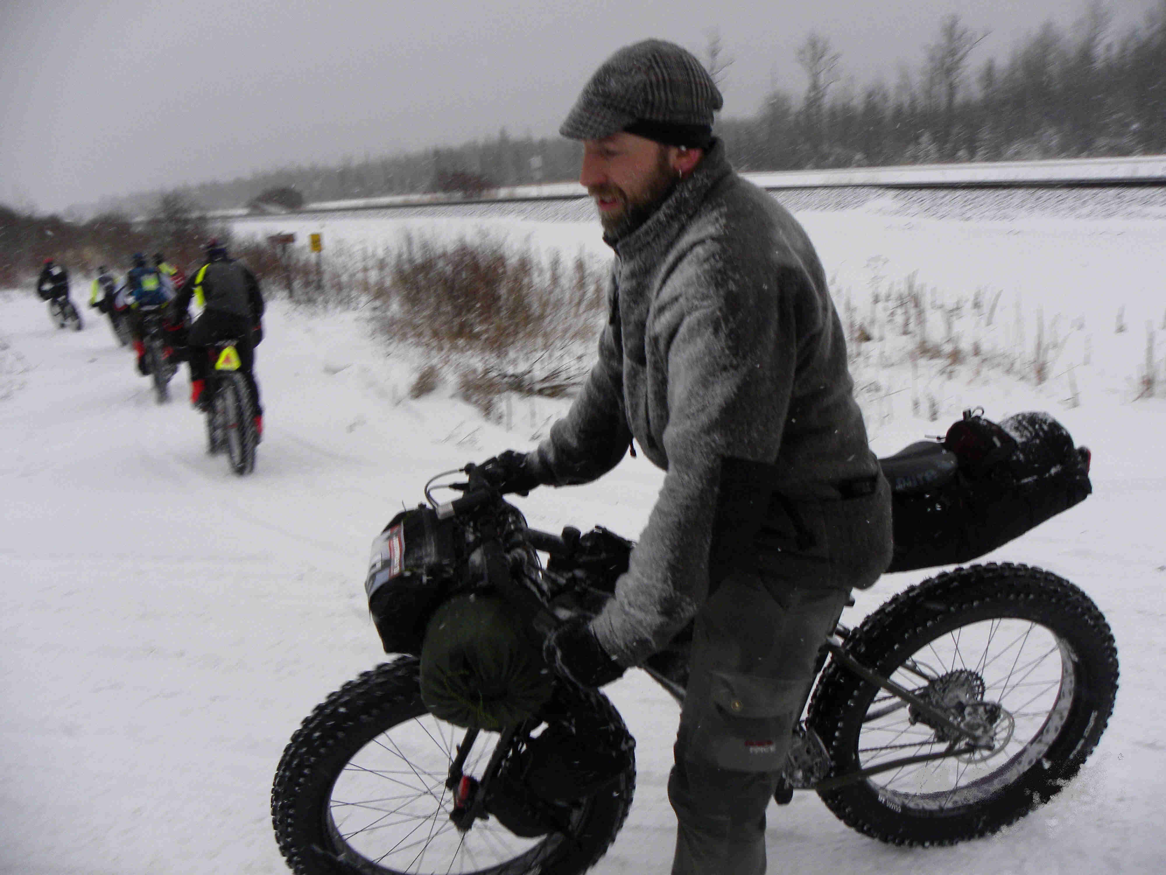Left side view of a cyclist wearing winter outerwear, on a Surly fat bike with gear, parked across a snowy trail
