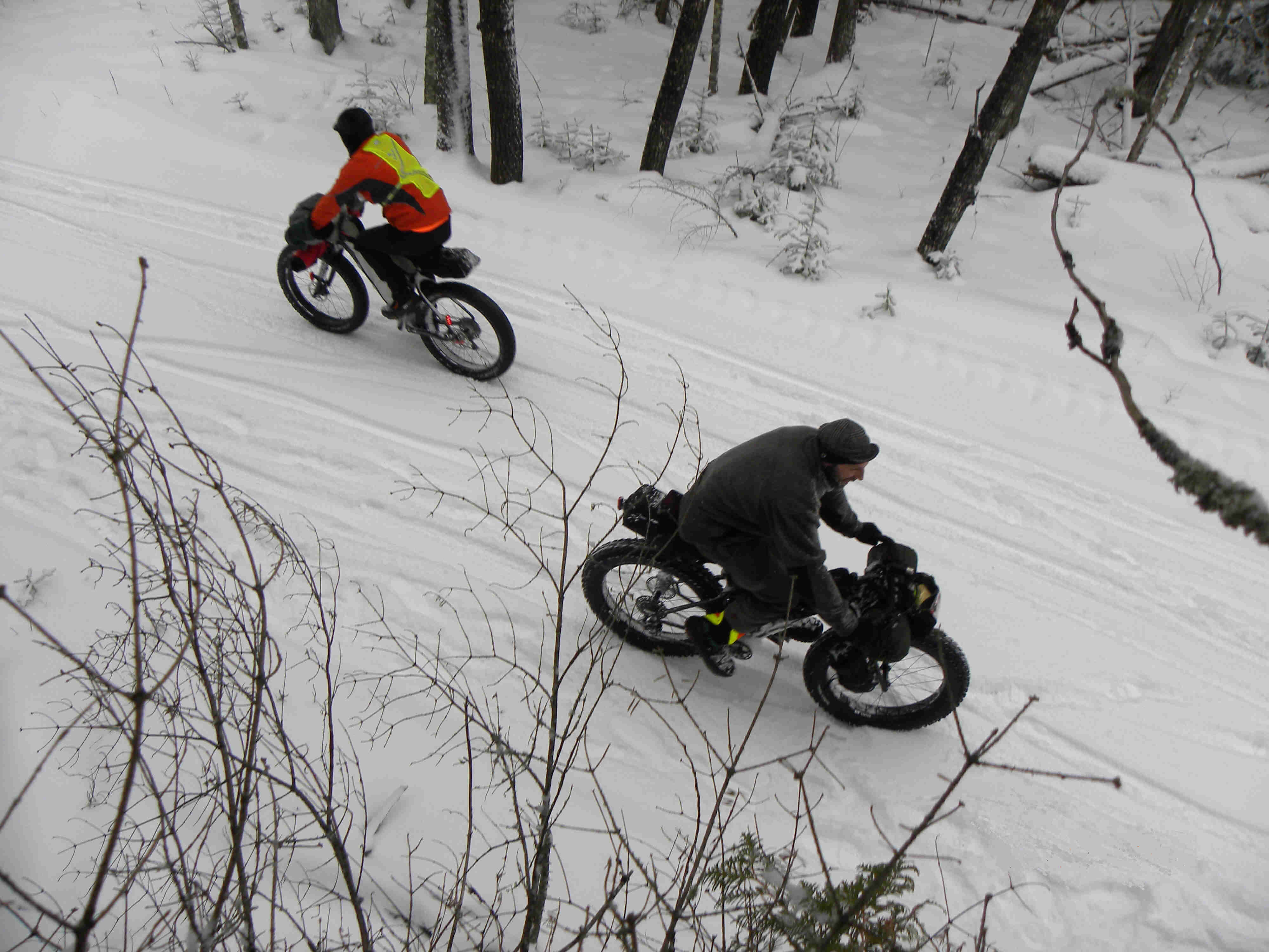 Downward view of 2 cyclists, wearing winter attire, riding their Surly fat bikes in opposite directions on a snowy trail