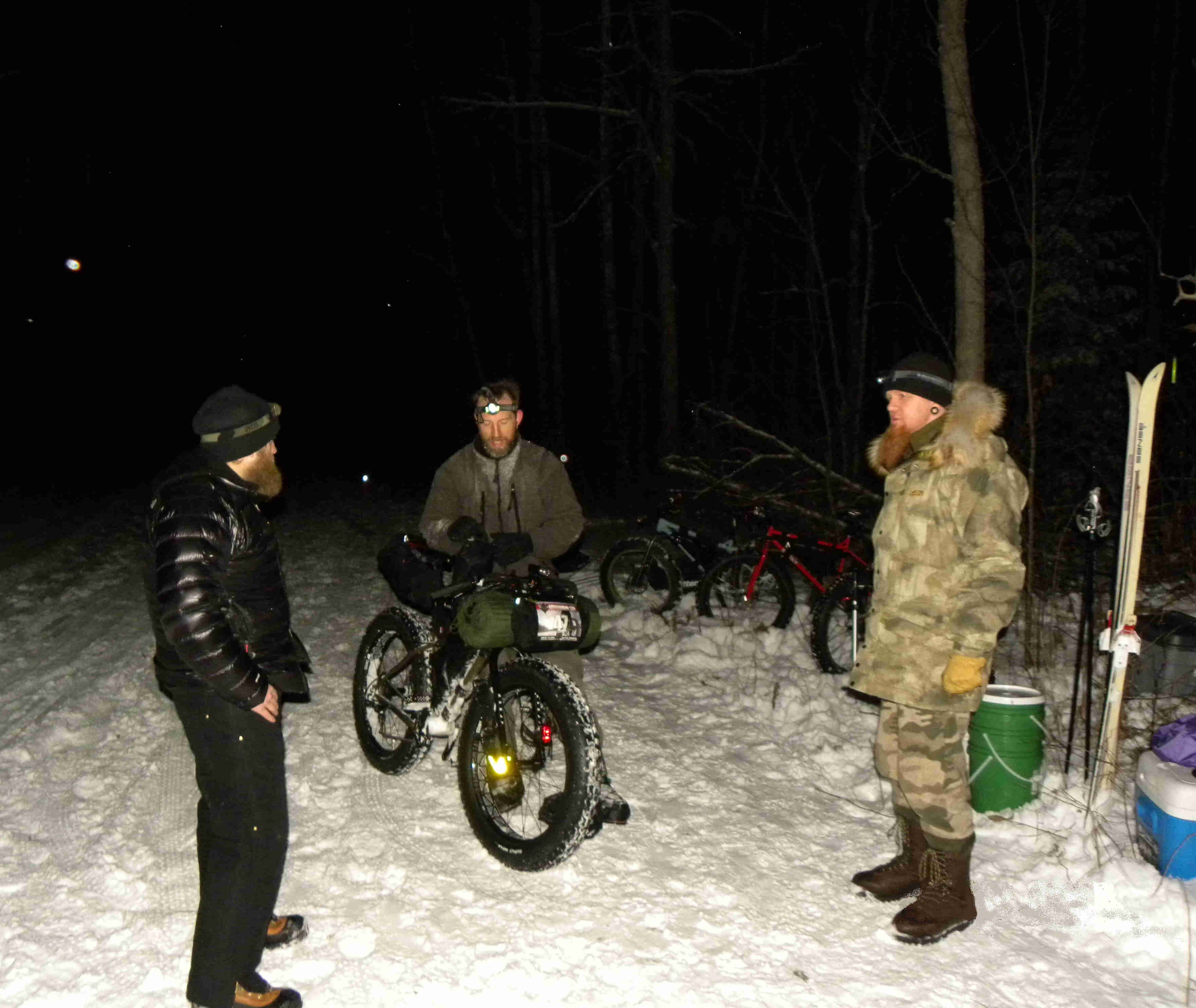 Two people wearing winter outerwear, with a cyclist and a Surly fat bike between them, on a snow covered road at night