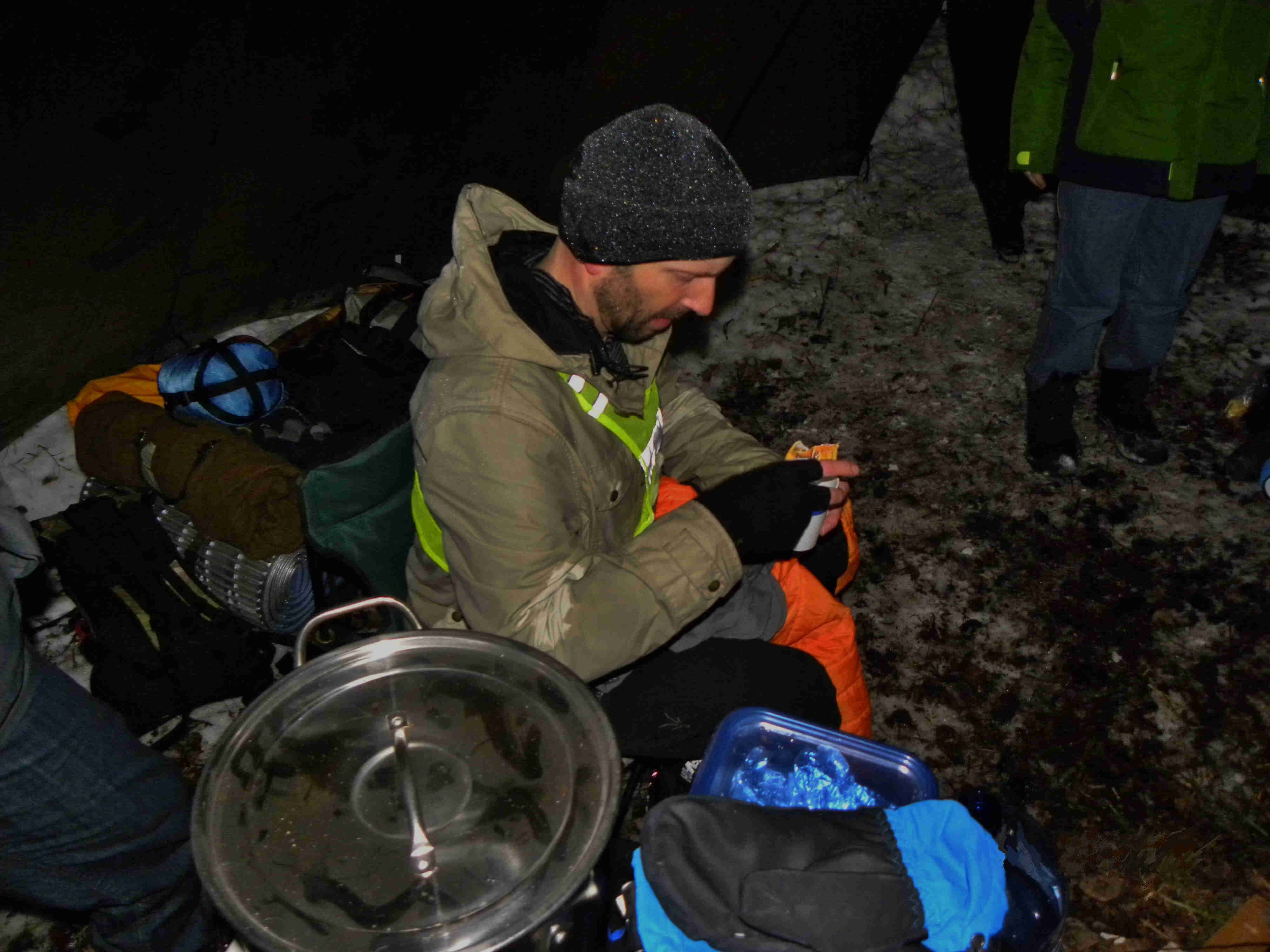 Downward view of a person, dressed in winter outerwear, sitting next to a pile of camp gear on the snowy ground at night