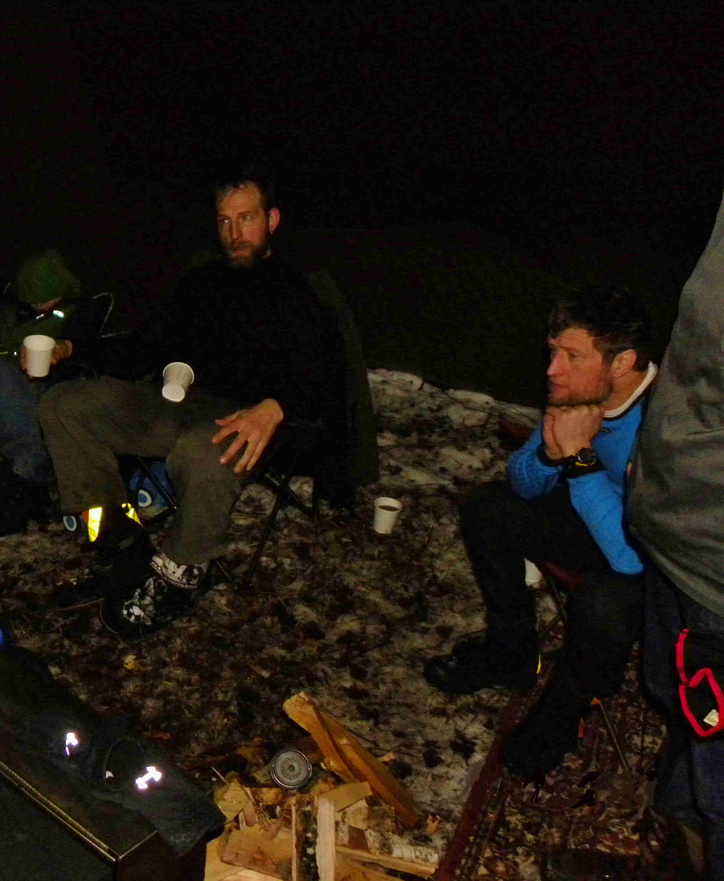 Front view of 2 people sitting in chairs, at a snowy campsite during the night 