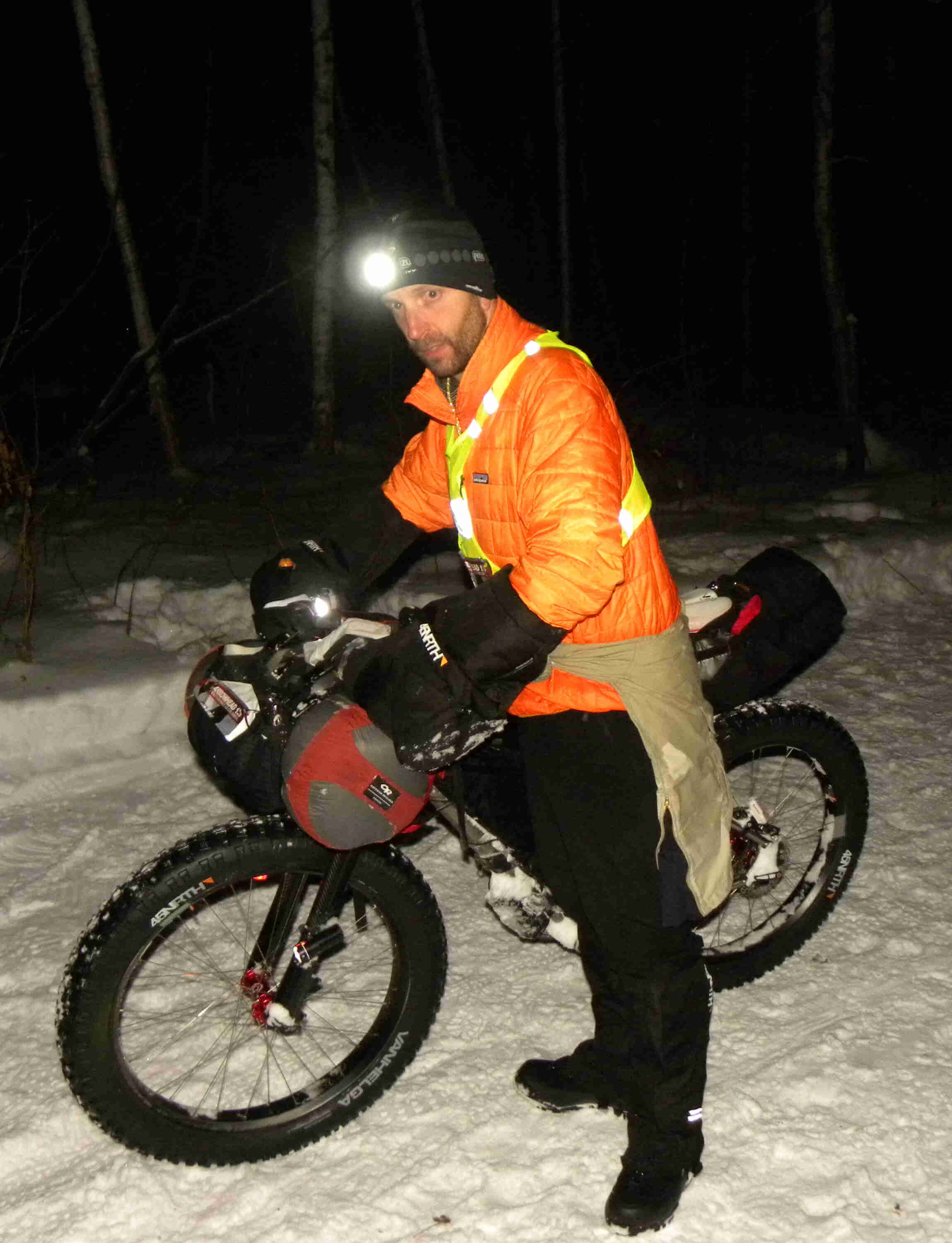 Right side view of a cyclist, wearing an orange coat, standing with their Surly fat bike in the snowy woods at night 