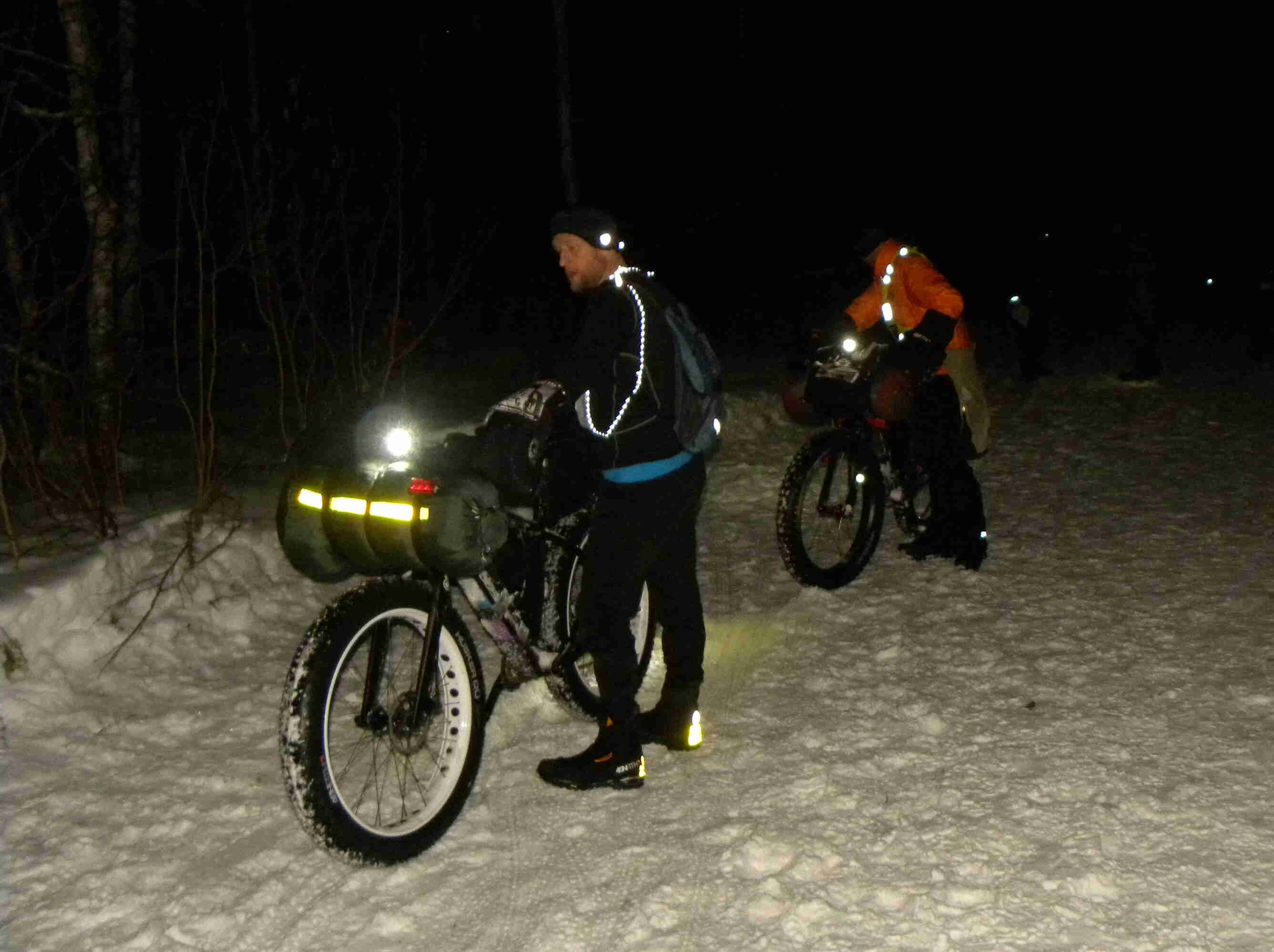 Front, left side view of 2 Surly fat bikes with cyclists standing on the left side, on a snowy trail at night