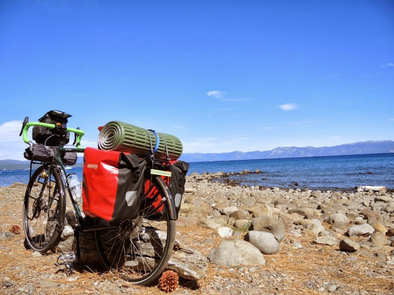 Rear, left side view of a Surly Disc Trucker bike, loaded with gear, on a rocky shore of a lake with mountains behind it