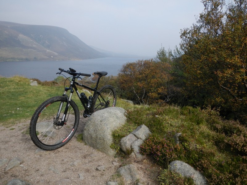 Front, left side view of a black Surly Karate Monkey bike, parked on a hilltop, with a lake and hills in the background