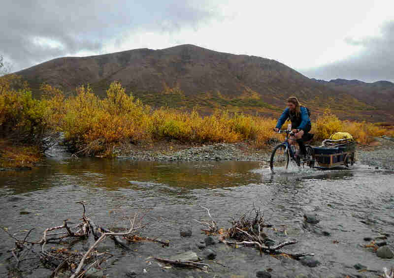 Left side view of a cyclist riding thru a stream, on a bike with trailer full of gear, with mountains in the background 