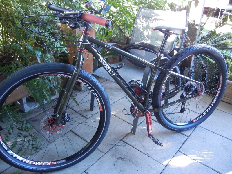 Left side view of a chocolate color Surly ECR bike, parked on a block patio against a chair, with shrubs behind it