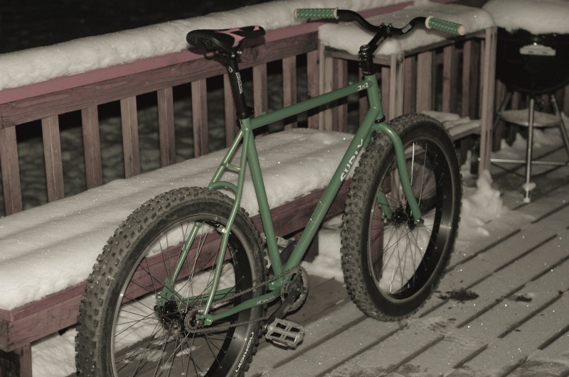 Right side view of a green Surly 1x1 bike, parked against a bench, on a snowy deck at night