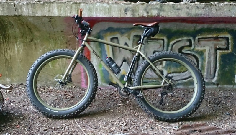 Left side view of an olive drab Surly Pugsley bike, parked on gravel, in front of a concrete barrier with graffiti on it