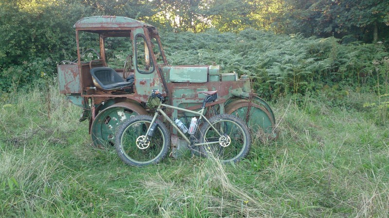 Left side view of an olive drab Surly bike, parked on the side of a rusty, broke down tractor, in a grass clearing