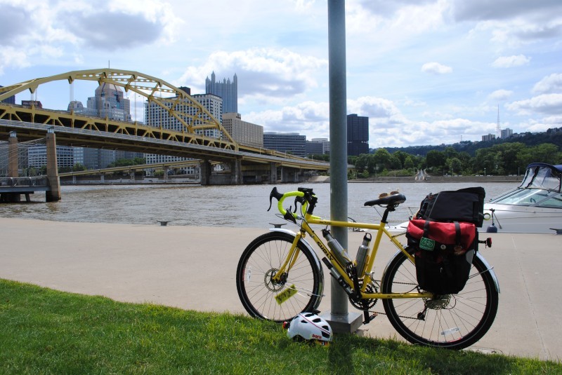 Left side view of a yellow Surly Disc Trucker bike, leaning on a light pole of a boardwalk, with a city bridge behind