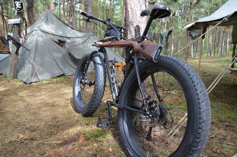 Rear, left side view of a black Surly fat bike with a rifle along the top tube, on a campsite with tents, in a forest