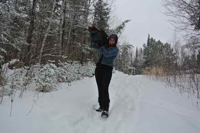 Front view of a person, holding gear up on their shoulder, on a snow covered trail with trees on the sides