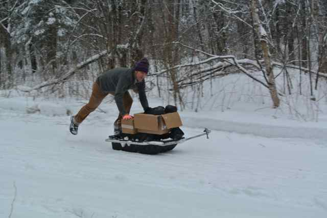Right side view of a person, pushing a sled with boxes on it, down a snow covered trail, with bare trees in background