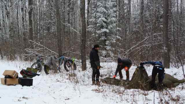 People setting up a tent in a snow covered clearing in the woods, with gear and bikes next to them
