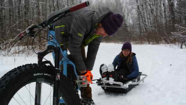 Front view of a cyclist, standing over a blue Surly fat bike, attaching a sled with a person in it, on a snowy trail