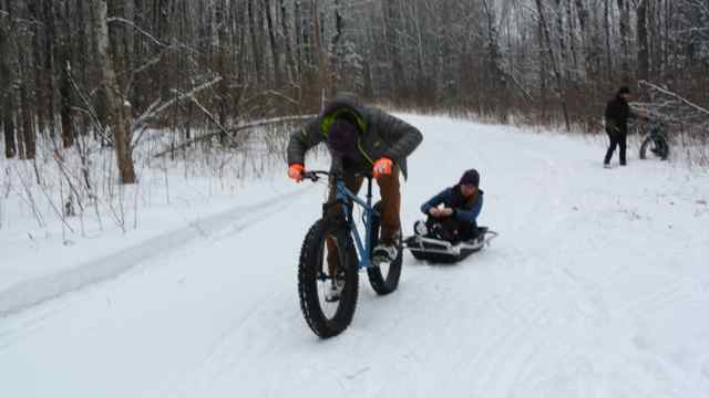 Front view of a cyclist, riding a blue Surly fat bike, pulling a person in a sled down a snowy trail in the woods