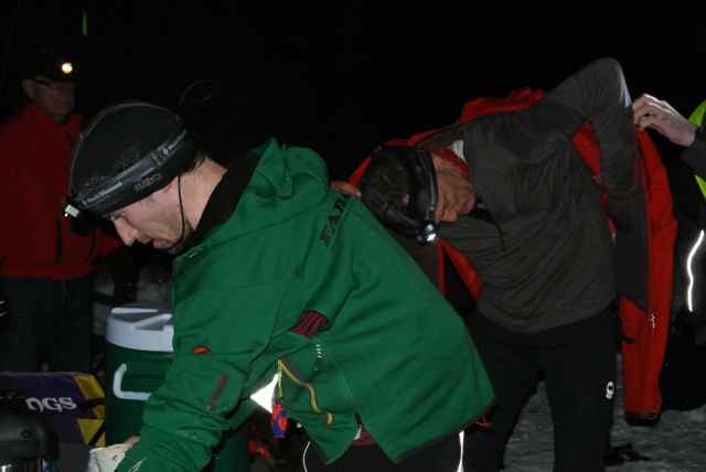 2 people dressed in winter outerwear and headlamps, at a campsite at night