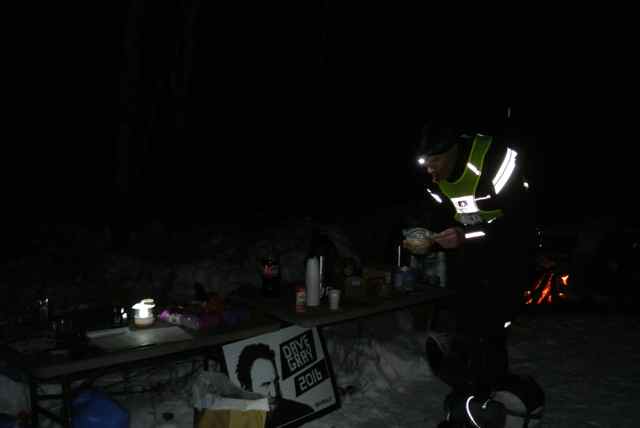 A person wearing winter outerwear and a reflective vest, standing in snow next to a table at night
