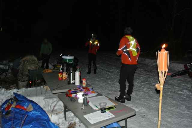Two people standing in snow, next to tables and gear, wearing winter outerwear and reflective jackets, at nighttime