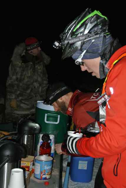 Right side view of a person wearing winter attire and a bike helmet, pouring a drink at night