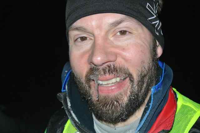 Headshot of a person, wearing a stocking cap, with an icicle hanging from their mustache, at night
