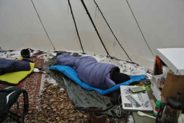 Downward view of a person, wrapped in a sleeping bag, on snowy ground inside a teepee tent