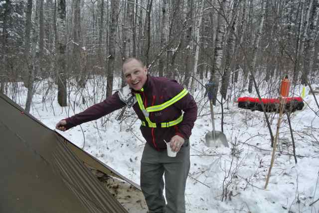 Front view of a smiling person, unzipping a teepee from the outside, with a snowy, bare woods in the background