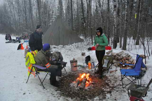 People wearing winter outerwear, standing and sitting around a campfire, with a tent behind, in the snowy woods