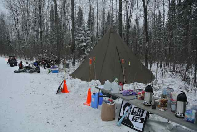 A table with food and beverages is next to a teepee tent, with fat bikes in the background, in the snowy woods