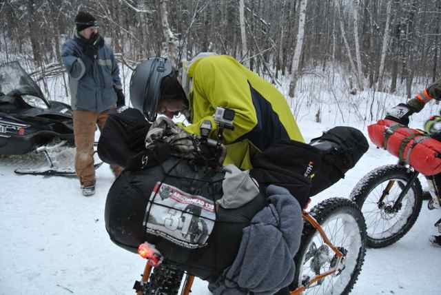 Front view of a cyclist dressed in winter outerwear, standing over a Surly fat bike loaded with gear, in the snowy woods