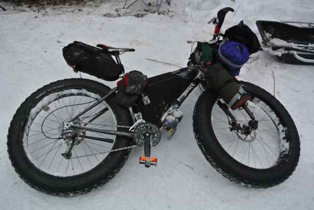 Downward, right side view of a Surly fat bike, loaded with gear, laying on the snow covered ground