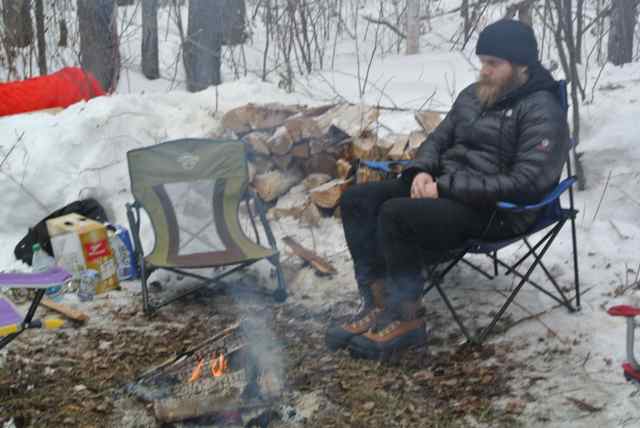 A person wearing winter attire, sits in a chair on snowy ground, on the right side of a campfire