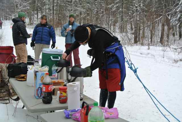A person winter outerwear, getting coffee from a thermos on a table, on a snow covered ground in the woods