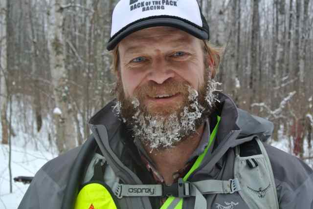 Head shot of a person with a frozen beard, wearing winter attire and a baseball cap, with the snowy woods behind them