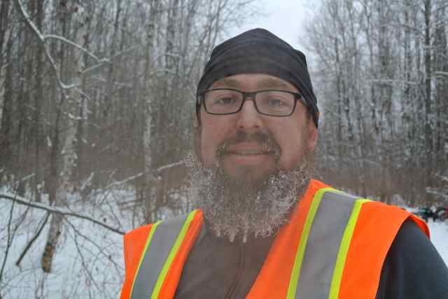 Head shot of a person with a frozen beard, wearing a reflective vest and glasses, with the snowy woods behind them