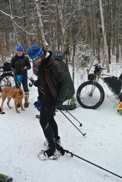 A person wearing winter outerwear, standing on snow, with a dog, Surly fat bikes and trees behind them