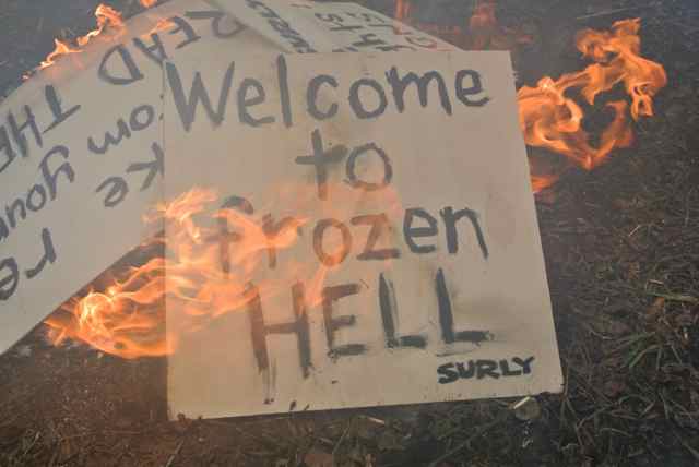 Downward view of a cardboard sign with the words, Welcome to Frozen Hell on it, burning in a campfire