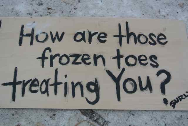 Downward view of a cardboard sign on the snow, with the words, How are those frozen toes treating you? on it