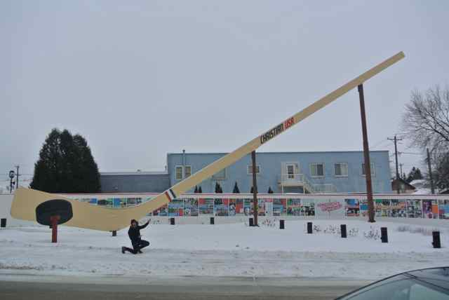A person kneeling down in the snow at the bottom of a giant hockey stick, with a light blue building in the background