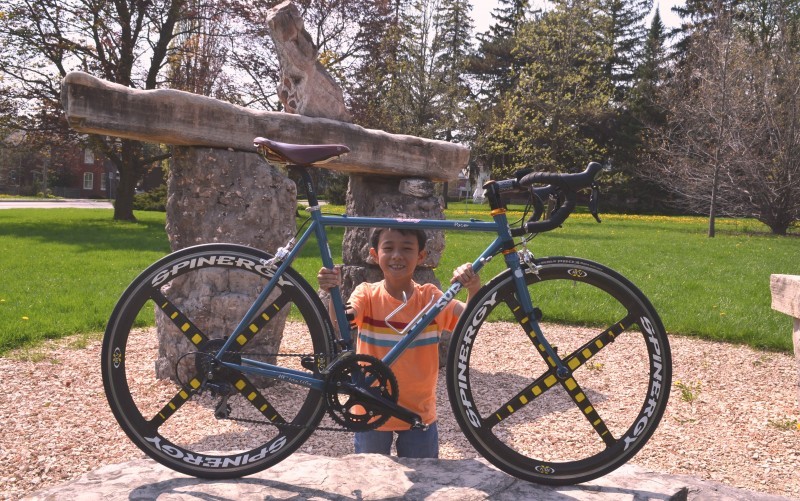 Right side view of a blue Surly bike, with a child standing behind holding the frame, with a stone statue behind them